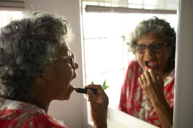 This image depicts a senior African American woman applying lipstick while looking in a mirror at home. It highlights themes of self-care, beauty routines, and personal grooming during quarantine and lockdown periods. This image can be used in articles or advertisements related to elderly care, beauty products for seniors, or mental well-being during isolation.