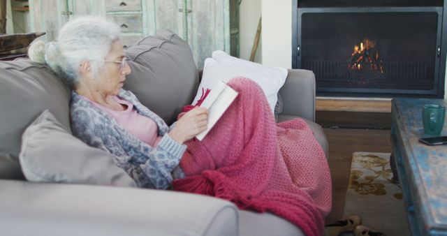 Elderly woman enjoying a book while sitting on a sofa covered with a warm pink blanket near a glowing fireplace. Ideal for lifestyle content, themes of relaxation and leisure, articles about hygge living, or promotions for retirement and leisurely activities for seniors.