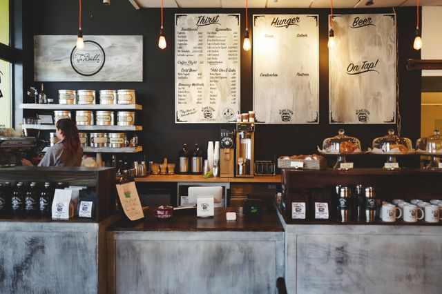 Cozy coffee shop with rustic decor featuring wooden counters, chalkboard menus, and hanging lights. A barista stands behind the counter, surrounded by jars, cups, and pastries. Perfect for illustrating a modern, casual coffee shop setting or showcasing small business ambiance.