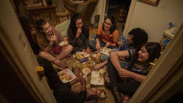 Young women sitting on rug sharing takeout and laughing in cozy apartment gathering