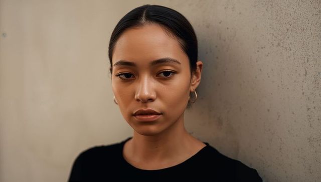 Pensive woman gazing downward against beige wall, moody portrait, gold hoop earrings