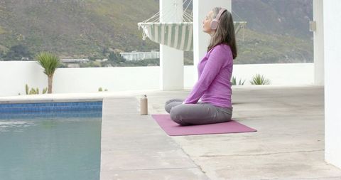 Mature woman meditating poolside on pink mat with headphones, terrace oasis and serenity