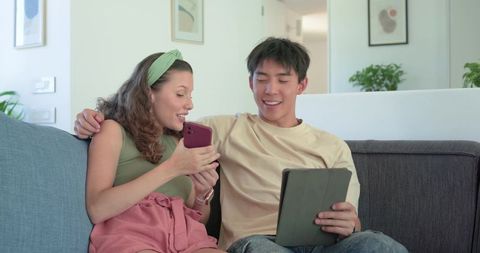 Young diverse couple sharing smartphone and tablet on sofa, modern cozy home lifestyle