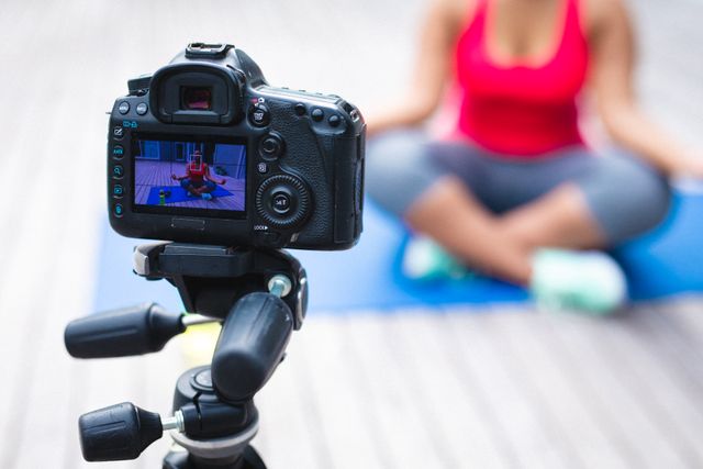 Female blogger recording a yoga session at home using a digital camera on a tripod. She is sitting on a yoga mat, wearing sportswear, and focusing on fitness and a healthy lifestyle. Ideal for content related to home workouts, fitness tutorials, wellness blogs, and online fitness classes.