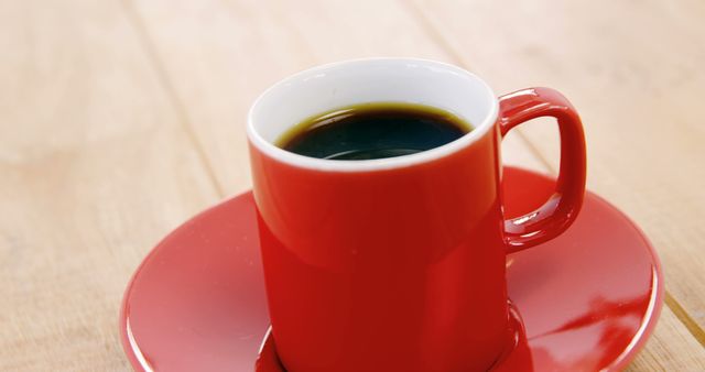 A red coffee cup filled with black coffee sits on a matching red saucer, placed on a wooden table, with copy space. Its vibrant color contrasts with the warm tones of the wood, suggesting a cozy coffee break atmosphere.