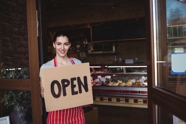 Female baker standing at the entrance of her bakery shop, holding an open sign and smiling. She is wearing a red and white striped apron, welcoming customers to her business. Ideal for use in articles about small businesses, local bakeries, entrepreneurship, and customer service.