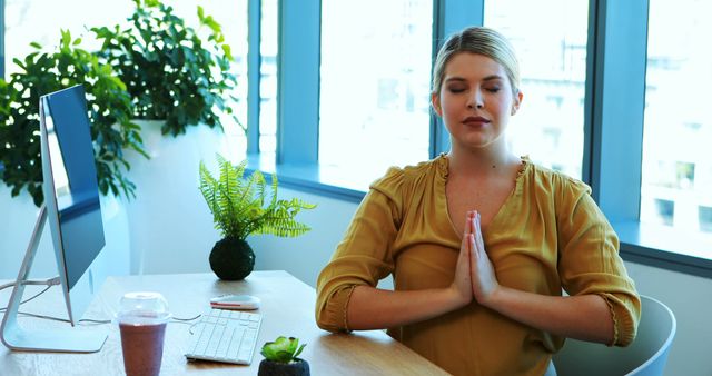 A young Caucasian woman is practicing meditation at her office desk, with copy space. Her serene posture and closed eyes suggest a moment of mindfulness amidst a busy work environment.