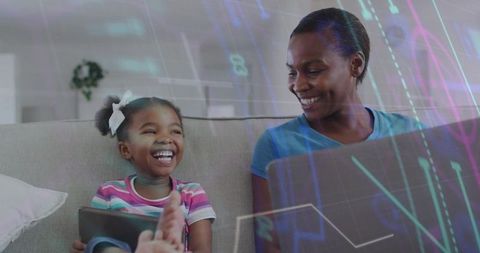 Laughing mother and daughter sharing tablet while using laptop on couch for digital learning