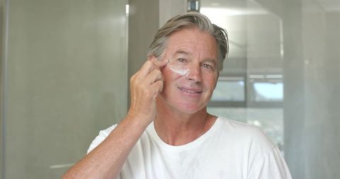 Mature man applying facial moisturizer at bathroom mirror during daily skincare routine