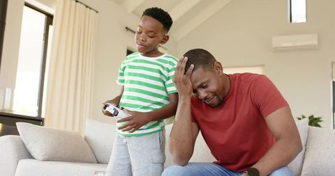 Tired father pausing while son plays video game on couch in bright modern living room