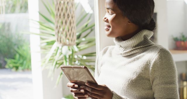 The woman stands near a sunlit window, holding a digital tablet and smiling while enjoying the natural light. This image can be used in marketing materials focusing on technology products, leisure time, comfortable home settings, or inclusive advertising campaigns.