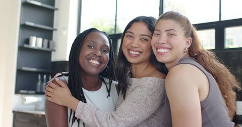 Diverse women hugging and smiling in sunlit modern kitchen, casual home friendship