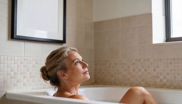 Middle-aged woman soaking in bathtub by window with mosaic tile border reflecting and relaxing