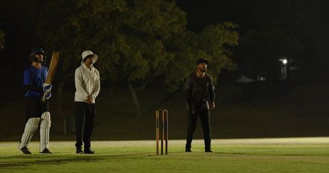 Night cricket players standing on floodlit park pitch awaiting ball, batter holding bat