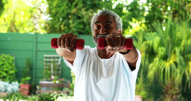 The senior man is lifting dumbbells in an outdoor garden. The greenery and sunny atmosphere suggest a healthy and active lifestyle. This image is ideal for promoting senior fitness, wellness products, articles on healthy aging, and fitness routines designed for elderly individuals.