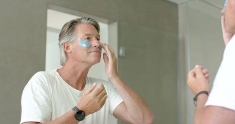 Mature man applying blue under-eye gel patches while checking mirror skincare routine