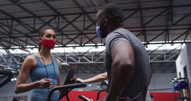 Personal trainer talking to gym client while both wearing masks. Useful for articles or advertisements about fitness training, gym environments, or health and safety practices during the pandemic.