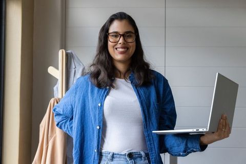 Confident woman smiling while holding laptop by window in bright modern home office