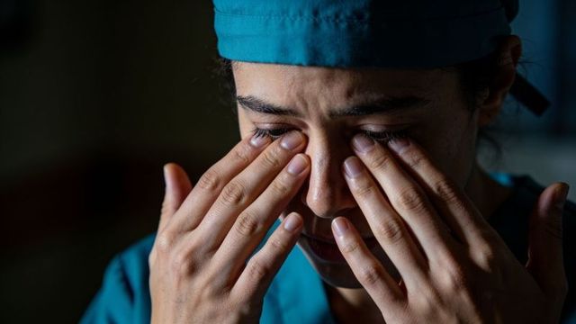 Tearful nurse rubbing eyes and pressing fingertips to face, conveying clinical fatigue