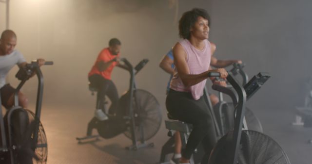 This image captures a diverse group cycling class in a modern gym setting. A woman in the foreground is actively cycling, focusing on her workout. Behind her, other participants are equally engaged in their exercise. The intensity of their efforts shows a commitment to fitness and a healthy lifestyle. Ideal for use in content related to group workouts, fitness programs, motivational fitness campaigns, gym promotions, and healthy lifestyle inspiration.