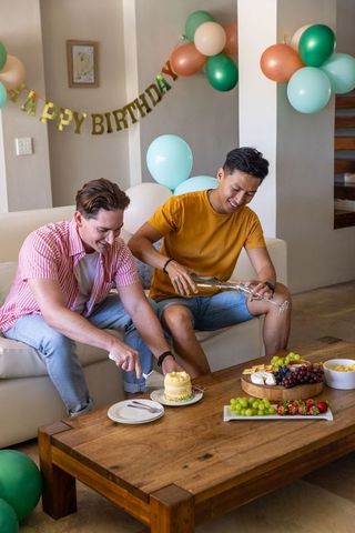 Diverse friends celebrating birthday at home cutting cake pouring bubbly on sofa