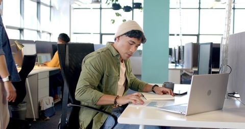 Young analyst reviewing charts at modern open-plan office desk with laptop in daylight