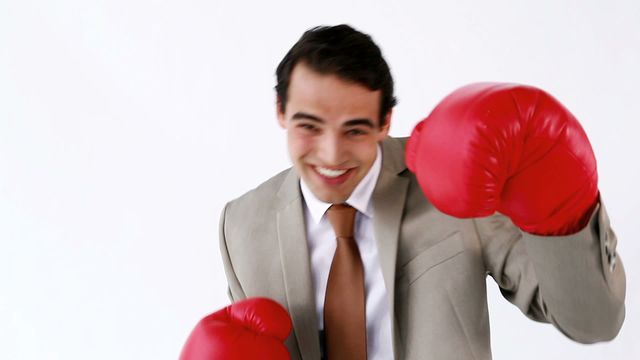 A businessman confidently smiles while wearing a professional suit and large red boxing gloves. The scene exudes a sense of humor and creativity, combining professionalism and sport. This can be used for themes related to confident leadership, blending corporate and playful elements, or promoting office wellness programs.