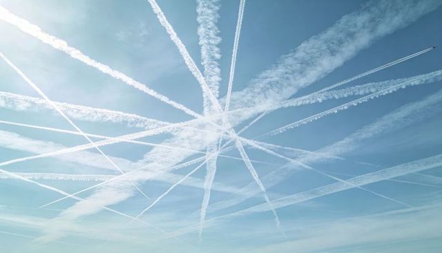 Contrails radiating across pale blue sky forming starburst pattern with jet trail