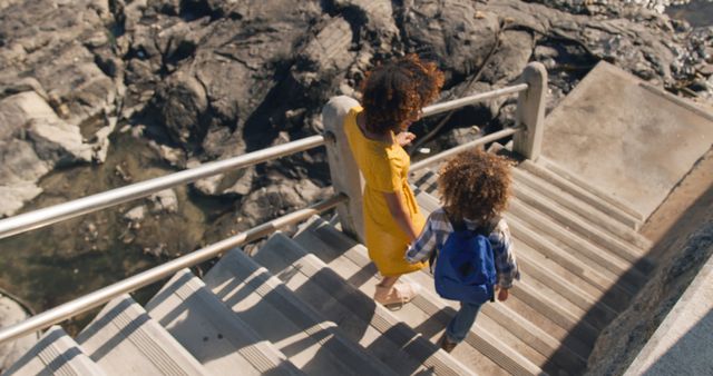 Happy biracial mother and son holding hands walking down steps to beach in the sun, copy space. Motherhood, childhood, togetherness, summer, vacations and free time, unaltered.