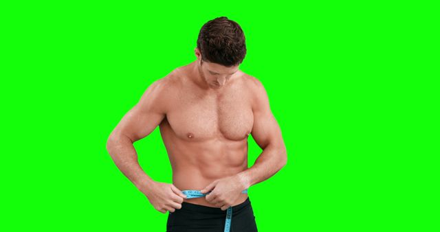 Fit man measures his waist using measuring tape against green screen background. Ideal for fitness programs, gym promotions, health articles, diet plans.