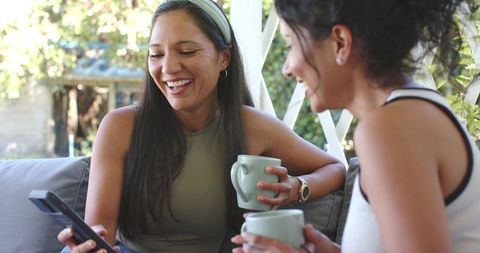 Smiling friends sharing coffee and smartphone on sunlit patio, casual outdoor morning chat