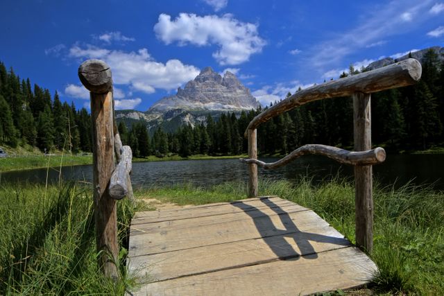 Peaceful landscape featuring alpine mountains under clear blue sky viewed from wooden bridge over lake surrounded by forest and grass. Perfect for promoting outdoor activities, hiking, adventure travel and nature retreats, bringing attention to scenic and tranquil vacation destinations.