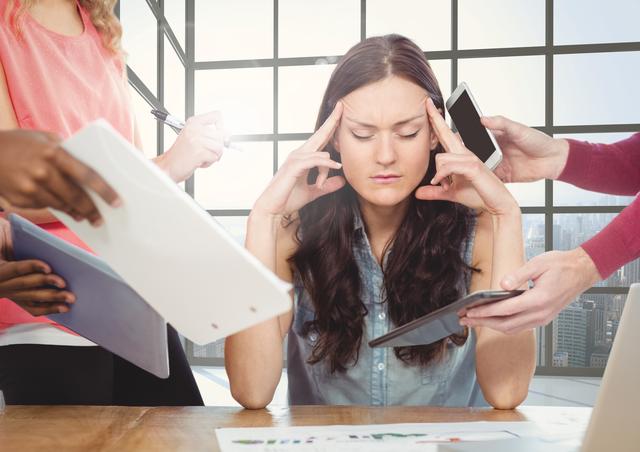 Businesswoman appearing stressed and overwhelmed is sitting at a desk in a modern office, surrounded by colleagues holding documents and electronic devices. This could be used to illustrate themes of workplace stress, multitasking, corporate pressure, and the demanding nature of professional environments.