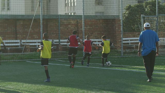 Multi-ethnic group of children engaged in soccer practice on a lush green football pitch on a sunny day. Two teams are seen actively playing, passing, and shooting the ball while a coach in a blue shirt watches over. Suitable for content related to youth sports, team activities, outdoor exercise, and coaching sessions.