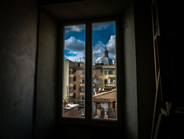 Scenic view of a historical city from inside a dark room through a window. The window frames an urban scene featuring historical buildings and a dome under a cloudy sky. Ideal for travel websites, architectural blogs, and promotional materials focused on urban tourism.