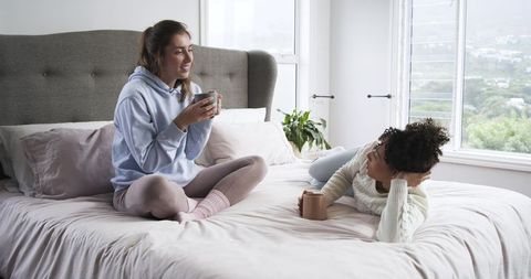 Cozy morning conversation between friends on bed sharing coffee in sunlit pastel bedroom