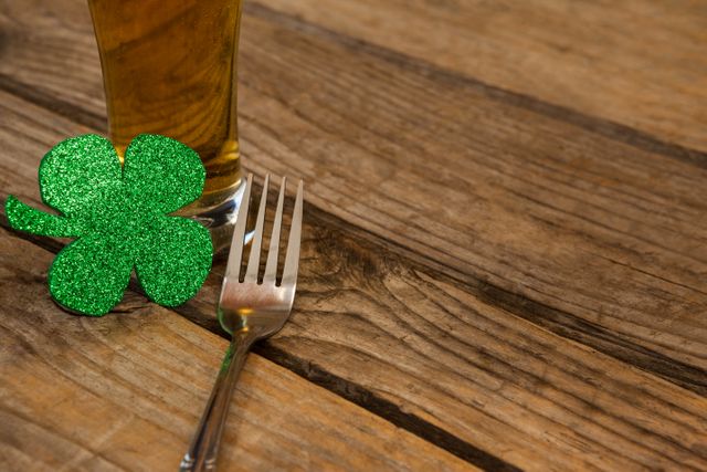 Perfect for illustrating St. Patrick's Day celebrations, this image features a glass of beer, a green glittery shamrock, and a fork on a wooden surface. Ideal for use in holiday promotions, festive event invitations, or social media posts celebrating Irish culture and traditions.