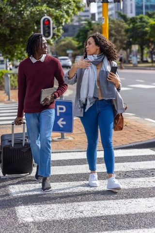 Young urban couple walking crosswalk pulling suitcase chatting while using phone and tablet