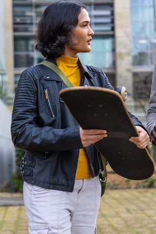 Woman holding skateboard and talking in urban plaza wearing leather jacket