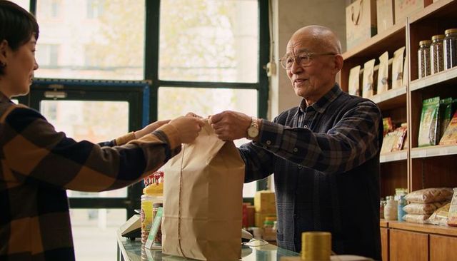 Senior shopkeeper handing brown paper bag to customer behind counter at corner grocery