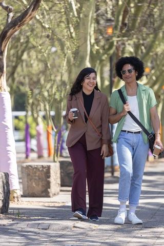 Diverse couple strolling on sunlit campus pathway holding takeaway coffees and smiling