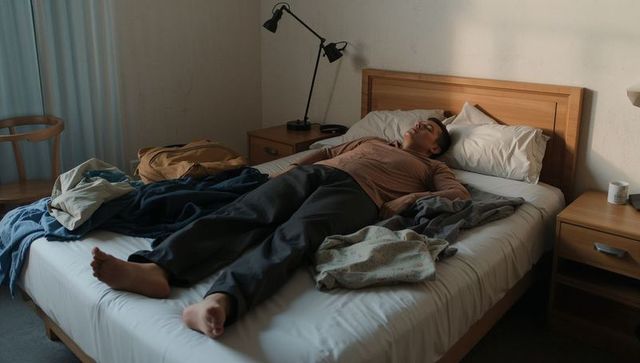 Young man resting on bed in minimalist bedroom with scattered clothes backpack and lamp