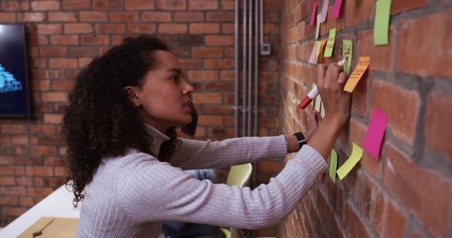 Biracial young professional sticking notes on brick wall. Wearing a striped sweater, her curly hair tied back, focusing on task