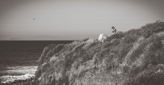 Individual sitting alone on a grassy coastal cliff, gazing at the ocean. Perfect for themes of solitude, contemplation, nature, reflection, relaxation, and quiet moments. Ideal for use in blogs, articles, mental health campaigns, environmental awareness projects, or travel promotions.