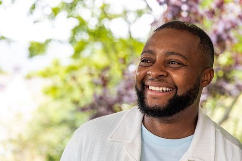 Joyful african man smiling and looking away wearing pale blue tee and jacket with green bokeh