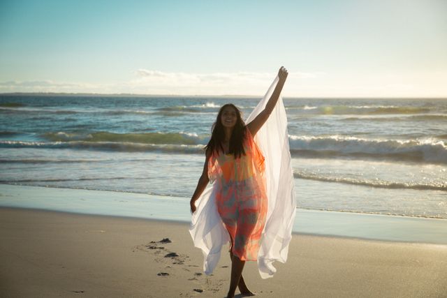 Happy biracial woman enjoying herself, dancing on beach holding scarf. holiday, freedom and leisure time at the beach.