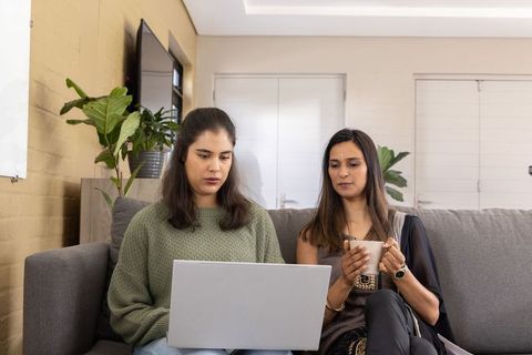 Two women collaborating on laptop while sitting on gray couch at home