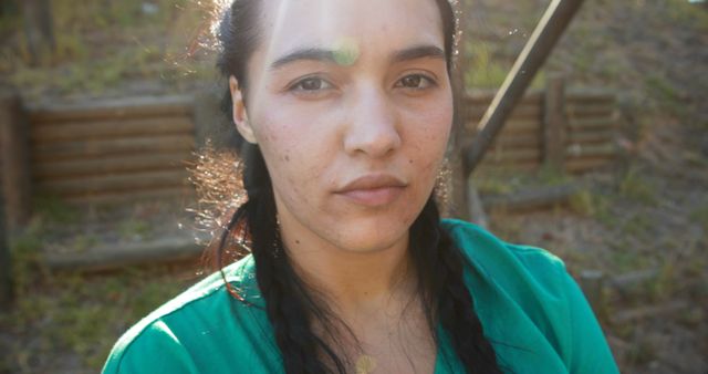 Young woman with braided hair standing outdoors in natural sunlight wearing a green shirt. She has a serious, pensive expression on her face. Suitable for articles on outdoor living, mental wellness, natural beauty, and lifestyle blogs or magazines.