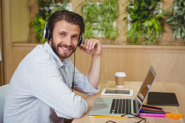 Smiling businessman wearing headphones and working on a laptop in a modern office with plants in the background. Ideal for use in articles about remote work, productivity, modern workspaces, and work-life balance.