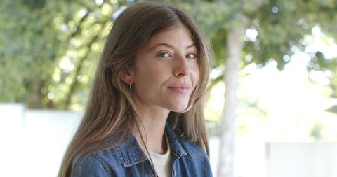 Smiling young woman standing in sunlit garden wearing denim jacket and hoop earrings